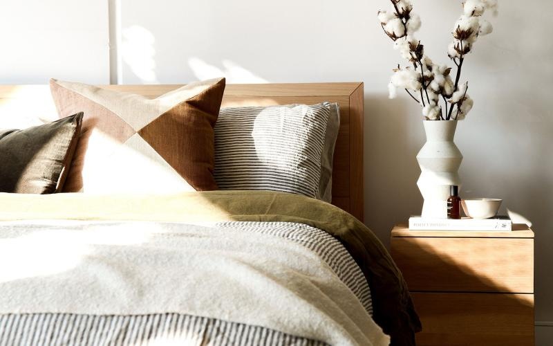 swath of sunlight shines across a well made bed and bedside table with a vase of cotton plants